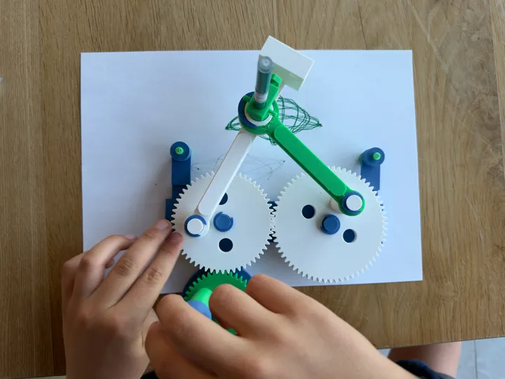 Hands turning the green crank while the linked arms draw a geometric pattern with a green pen.