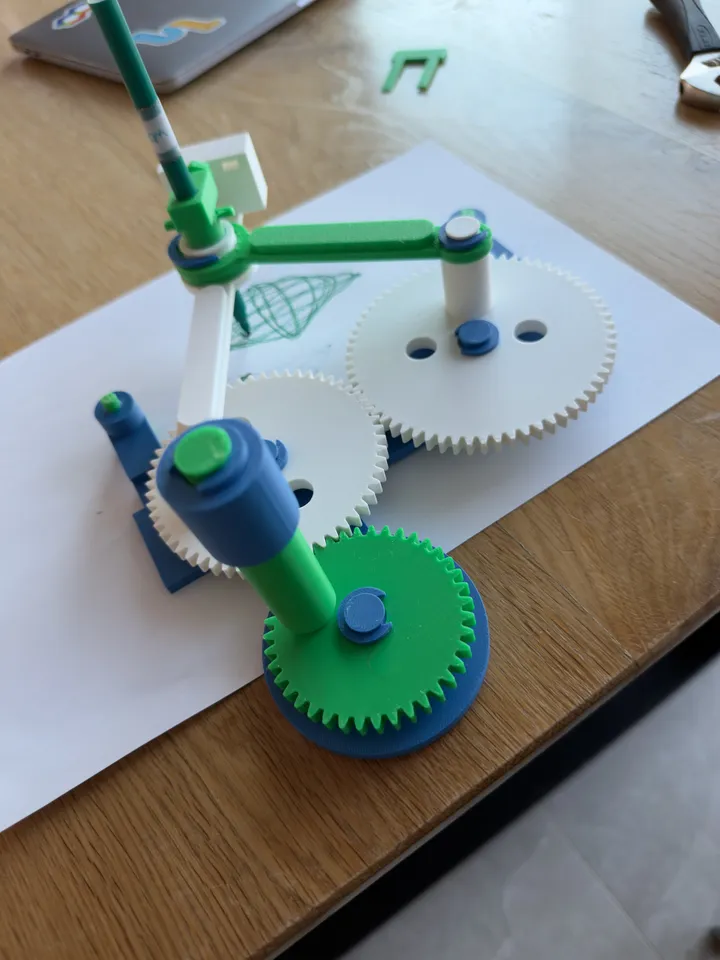 The 3D-printed drawing machine, with green and white gears and arms holding a pen over a geometric drawing.