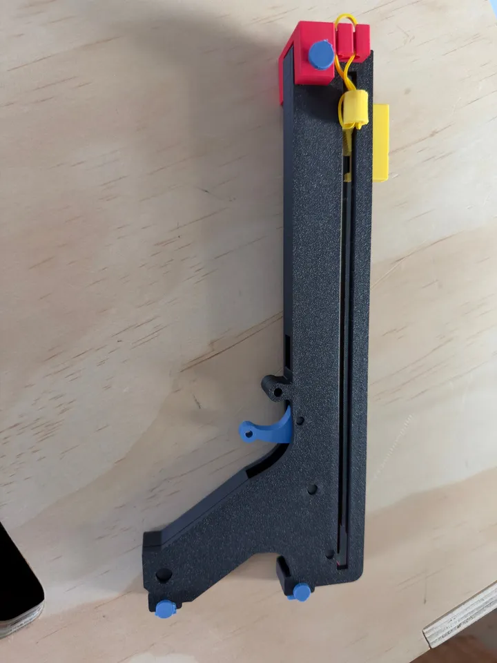 The 3D-printed paper-plane launcher on a wooden table, with a black body, blue trigger and fasteners, and red and yellow parts holding the elastic bands.